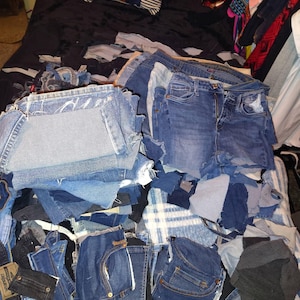 May include: A large collection of assorted denim fabric scraps and cut-up blue jeans pieces in various shades of blue, indigo, and black. The pile includes denim pockets, waistbands, and frayed fabric segments of different sizes and textures.
