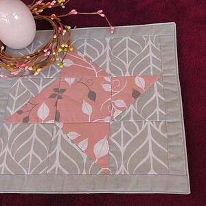 Peut inclure: Un chemin de table matelassé gris et rose avec un motif de feuilles et un motif en forme d'étoile au centre.
