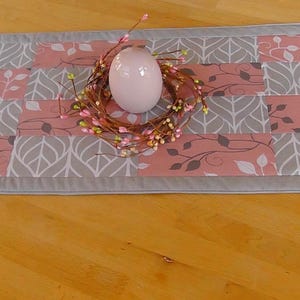 Könnte beinhalten: Ein grauer und rosa Tischläufer mit einem Blatt-Muster. Der Läufer ist mit einem weißen Ei und einem Kranz aus Zweigen und rosa Blüten verziert.