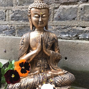 May include: A gold-colored Buddha statue sitting in a meditative pose with hands clasped in front of the chest. The statue is decorated with intricate details and has a small candle holder at the base.