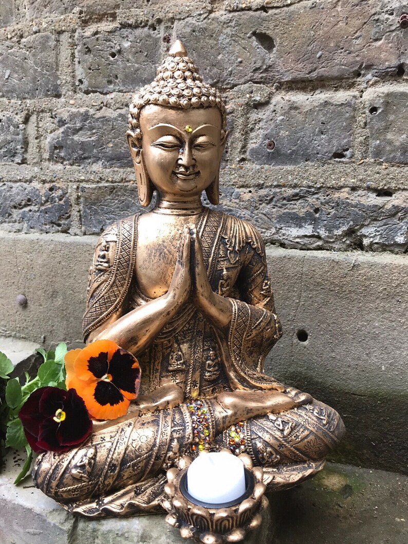 May include: A gold-colored Buddha statue sitting in a meditative pose with hands clasped in front of the chest. The statue is decorated with intricate details and has a small candle holder at the base.