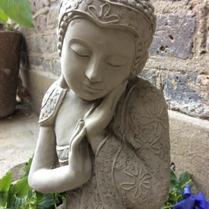 May include: A gray stone statue of a meditating Buddha with closed eyes and hands clasped in front of the face. The statue is decorated with intricate floral patterns and is sitting on a bed of green leaves and blue flowers.