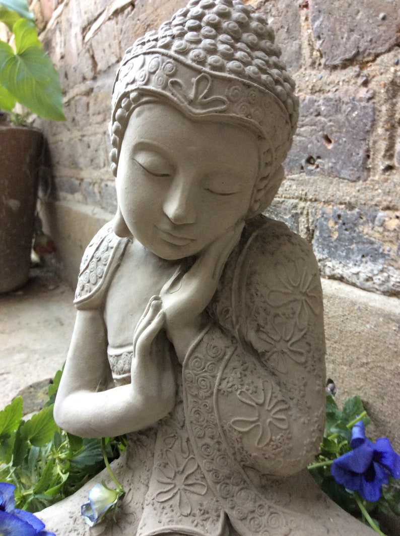 May include: A gray stone statue of a meditating Buddha with closed eyes and hands clasped in front of the face. The statue is decorated with intricate floral patterns and is sitting on a bed of green leaves and blue flowers.