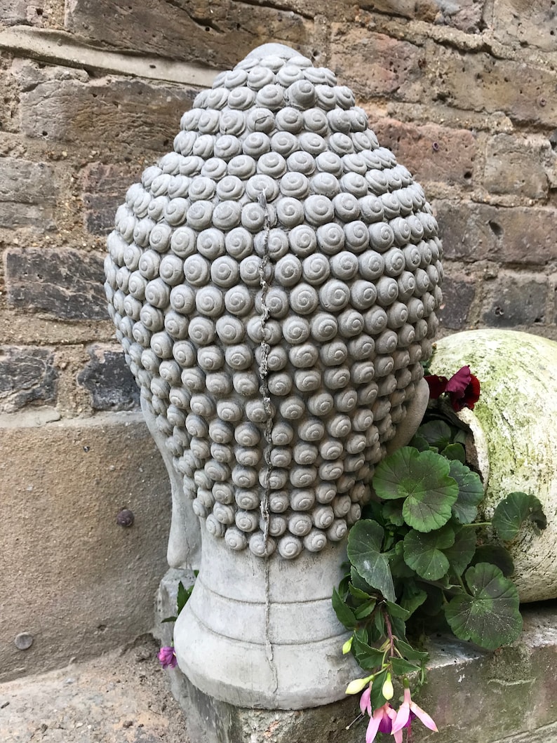 May include: A gray concrete Buddha head sculpture with a detailed, textured design. The head is placed on a stone surface with green plants and pink flowers growing around it.