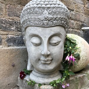 May include: A gray stone Buddha head sculpture with closed eyes and a serene expression. The sculpture is decorated with intricate details and is surrounded by green foliage and pink flowers.