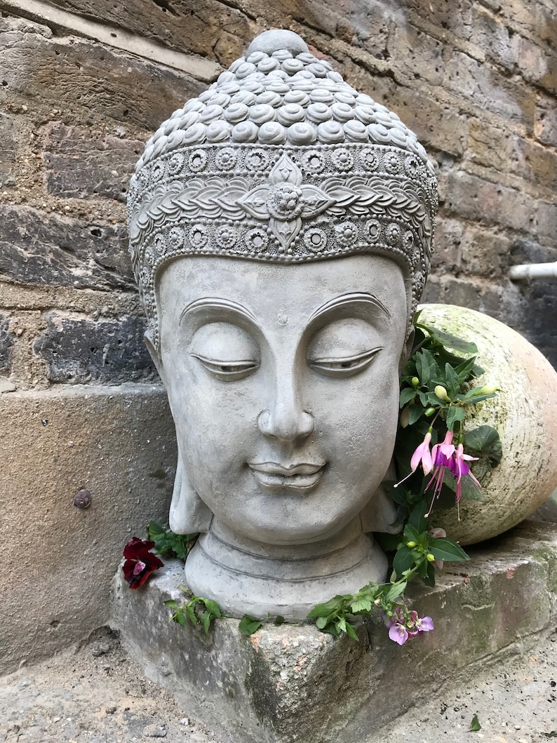 May include: A gray stone Buddha head sculpture with closed eyes and a serene expression. The sculpture is decorated with intricate details and is surrounded by green foliage and pink flowers.