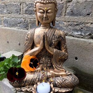 May include: A gold-colored Buddha statue sitting in a meditative pose with hands clasped in front. The statue is decorated with intricate details and has a small candle holder at the base.