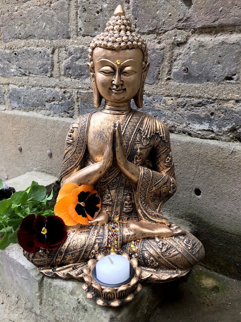 May include: A gold-colored Buddha statue sitting in a meditative pose with hands clasped in front. The statue is decorated with intricate details and has a small candle holder at the base.