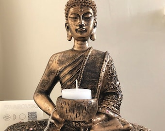 May include: A gold-toned Buddha statue with intricate details sits in a meditative pose, holding a small bowl with a white candle inside. The statue is decorated with small pink rhinestones.
