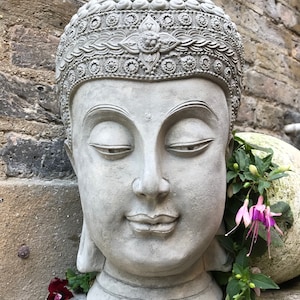 May include: A gray stone Buddha head sculpture with intricate details on the crown. The sculpture is placed on a stone surface with green plants and purple flowers.