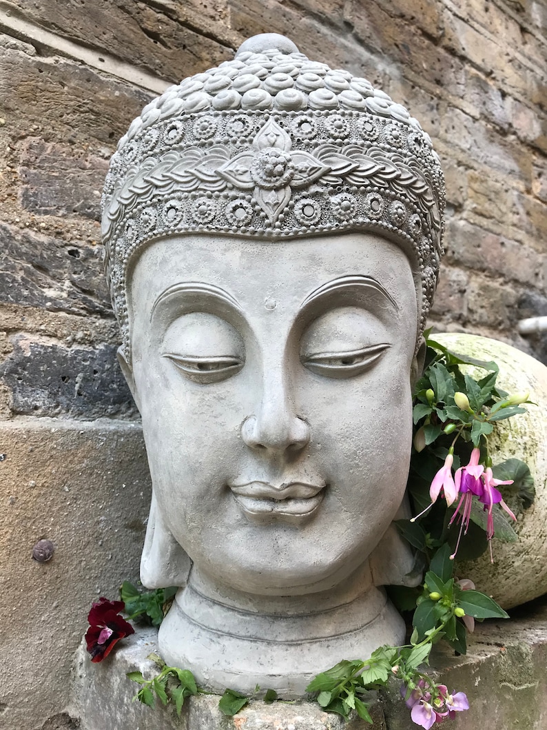 May include: A gray stone Buddha head sculpture with intricate details on the crown. The sculpture is placed on a stone surface with green plants and purple flowers.