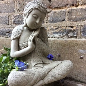 May include: A gray stone statue of a meditating Buddha with intricate details and a serene expression. The statue is sitting on a stone surface with a few purple flowers in the foreground.