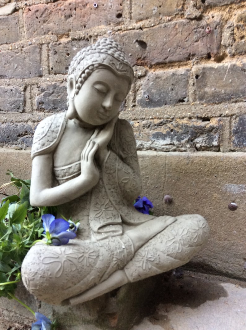 May include: A gray stone statue of a meditating Buddha with intricate details and a serene expression. The statue is sitting on a stone surface with a few purple flowers in the foreground.