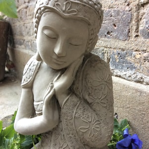 May include: A gray stone statue of a meditating Buddha with intricate floral designs on the robe. The statue is sitting with its head resting on its hand.