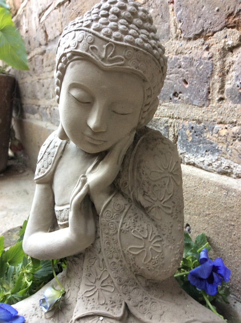 May include: A gray stone statue of a meditating Buddha with intricate floral designs on the robe. The statue is sitting with its head resting on its hand.
