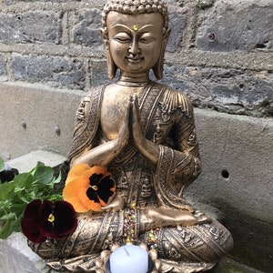 May include: A gold-colored Buddha statue sitting in a meditative pose with hands clasped in front of the chest. The statue is decorated with intricate details and has a small candle holder in front of it.