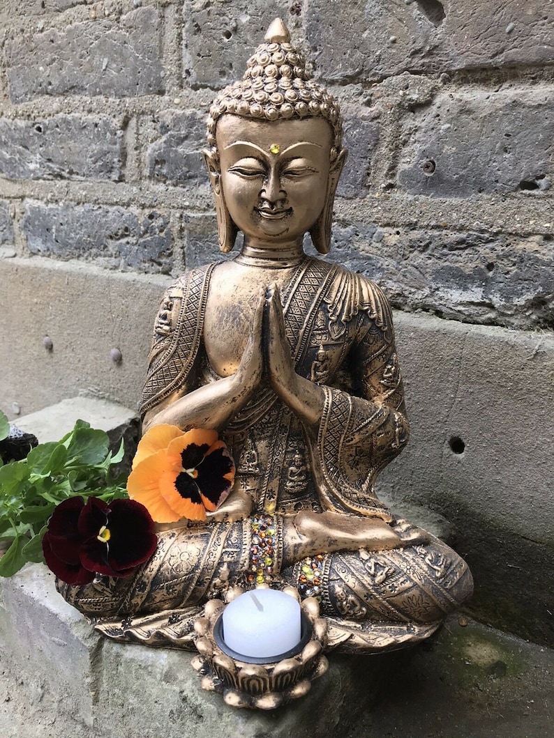 May include: A gold-colored Buddha statue sitting in a meditative pose with hands clasped in front of the chest. The statue is decorated with intricate details and has a small candle holder in front of it.