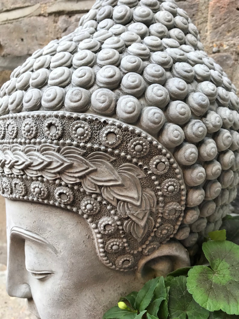 May include: A gray concrete Buddha head planter with intricate details and a floral design. The planter is partially filled with green plants.