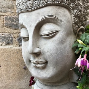 May include: A gray stone Buddha head planter with a detailed crown and closed eyes. The planter is decorated with green and purple flowers.