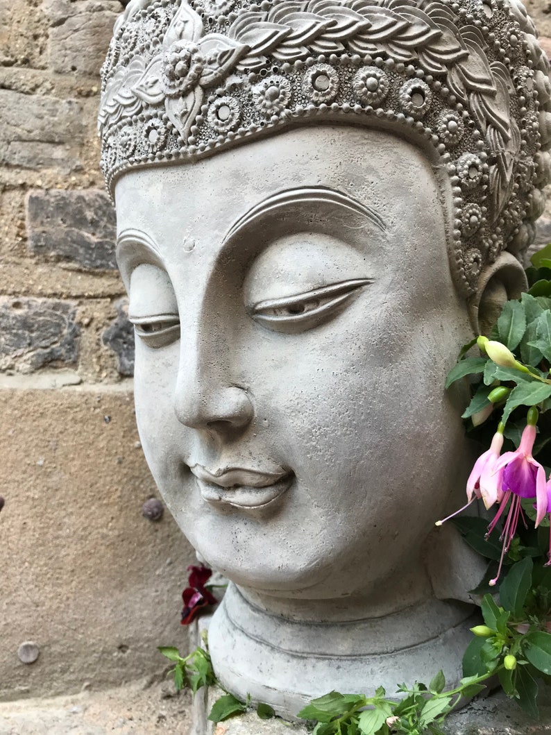 May include: A gray stone Buddha head planter with a detailed crown and closed eyes. The planter is decorated with green and purple flowers.