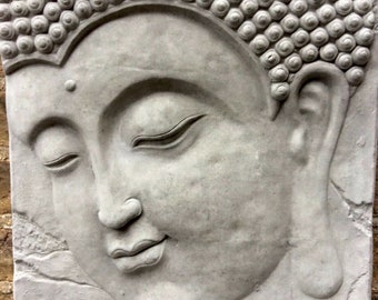 May include: A gray, rectangular Buddha face sculpture with closed eyes and a serene expression. The sculpture has detailed hair and is mounted on a brick wall. The sculpture is made of a stone-like material.