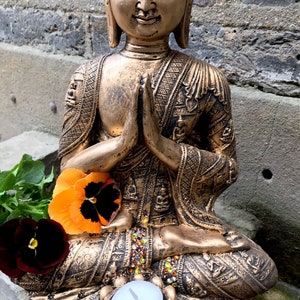 May include: A gold-colored Buddha statue in a seated meditation pose with hands clasped in prayer. The statue is decorated with intricate details and has a small candle holder at the base.