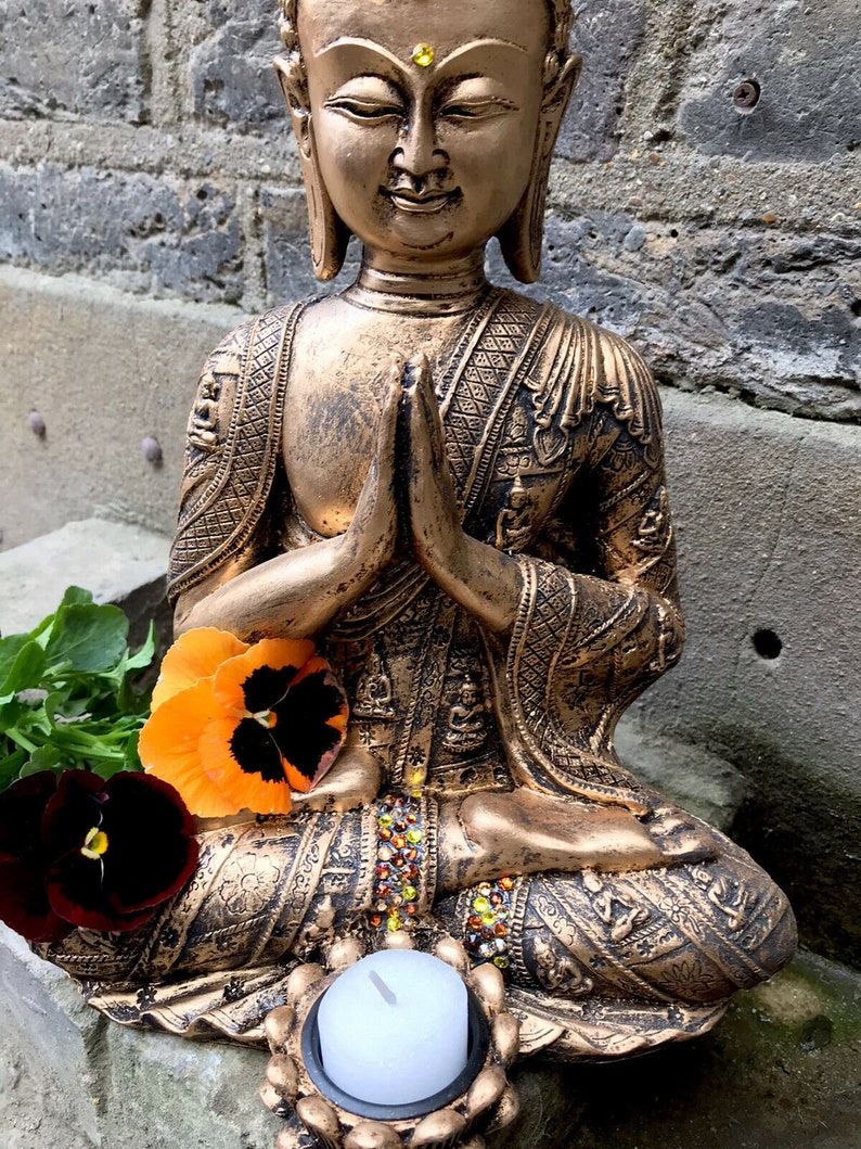 May include: A gold-colored Buddha statue in a seated meditation pose with hands clasped in prayer. The statue is decorated with intricate details and has a small candle holder at the base.