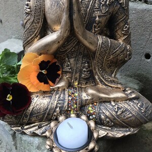 May include: A gold-toned Buddha statue in a seated meditation pose with a small white candle in a decorative holder. The statue is adorned with intricate details and has a jeweled design around the base.