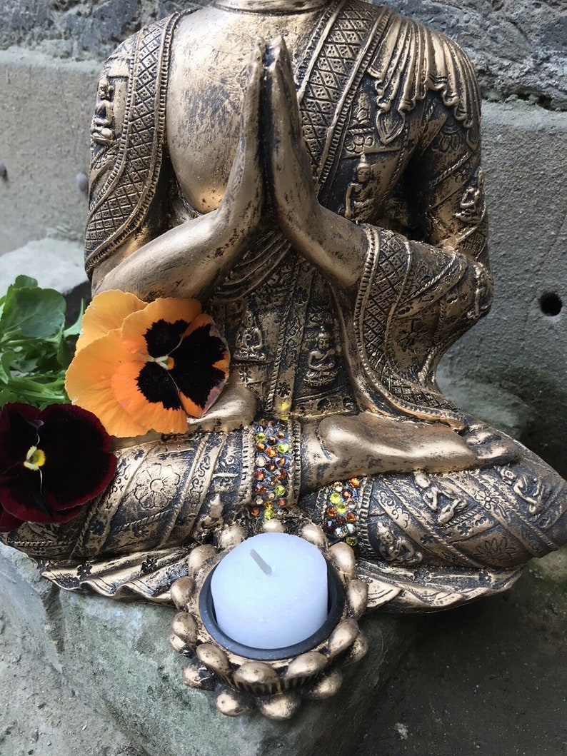 May include: A gold-toned Buddha statue in a seated meditation pose with a small white candle in a decorative holder. The statue is adorned with intricate details and has a jeweled design around the base.