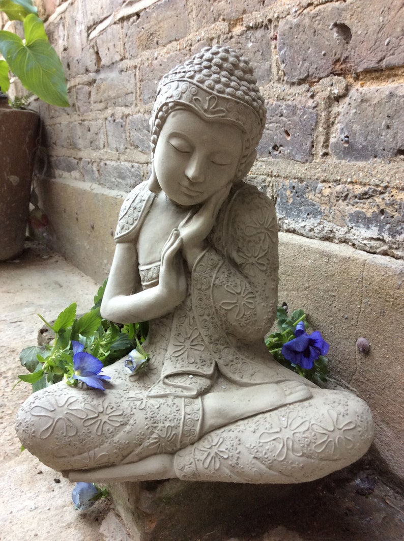 May include: A gray stone statue of a meditating Buddha with intricate details and a serene expression. The statue is surrounded by green plants and blue flowers.