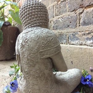 May include: A gray stone Buddha statue with intricate floral designs sits in a meditative pose. The statue is placed on a brick surface with a few purple flowers in the foreground.