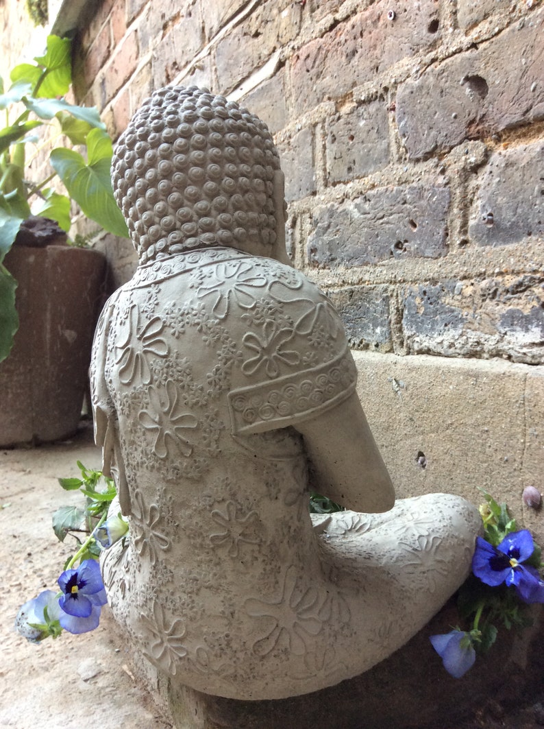 May include: A gray stone Buddha statue with intricate floral designs sits in a meditative pose. The statue is placed on a brick surface with a few purple flowers in the foreground.