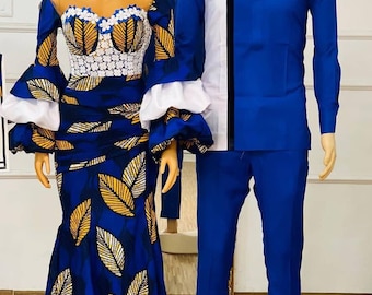 Op de afbeelding: Een bijpassende outfitset voor een stel, in een blauw-gouden kleurenschema. De jurk van de vrouw heeft een aansluitend lijfje met een witte kanten overlay en een zwierige rok met een bladmotief. De outfit van de man omvat een ​​lang mouw shirt met een wit paneel en een bijpassende broek.