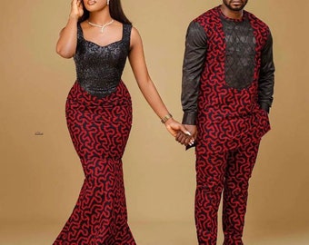 May include: A couple wearing matching red and black patterned outfits. The woman is wearing a sleeveless dress with a black bodice and a red and black patterned skirt. The man is wearing a long-sleeved shirt and pants with the same pattern. They are holding hands and smiling.