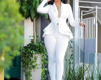 May include: A white pantsuit with a fitted jacket and flared peplum detail. The outfit includes matching slim-fit pants. The person is wearing clear heels and jewelry. The background includes greenery and a modern building.