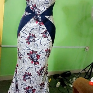 May include: A white and blue floral print dress with a navy blue sash and a one-shoulder neckline. The dress has a fitted bodice and a flowing skirt.