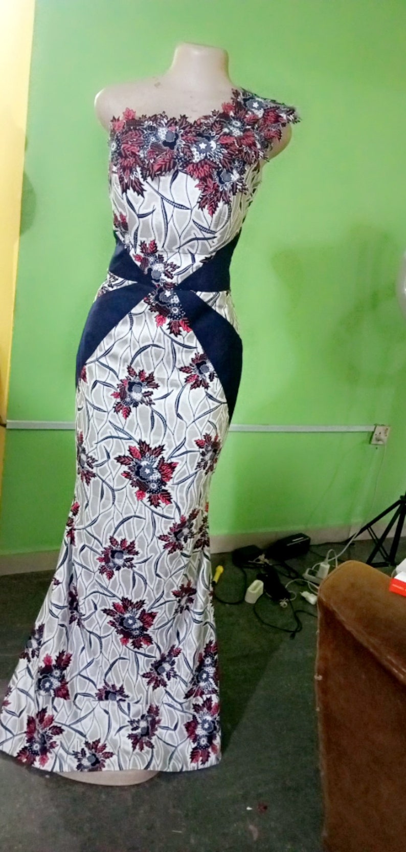 May include: A white and blue floral print dress with a navy blue sash and a one-shoulder neckline. The dress has a fitted bodice and a flowing skirt.