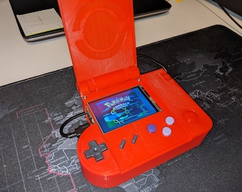 Op de afbeelding: Een felrode handheld gameconsole met een scherm waarop "Pokémon Emerald Version" wordt weergegeven. Het apparaat heeft een navigatiepad, knoppen en een scharnierende bovenkant. Een zwarte USB-kabel is aangesloten.