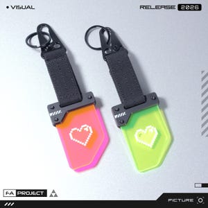 Op de afbeelding: Twee sleutelhangers met een pixelhartontwerp. Eén is roze, de andere neon groen. Elk heeft een zwarte band en een zwarte metalen sluiting. De tekst "RELEASE 2026" is rechtsboven zichtbaar.