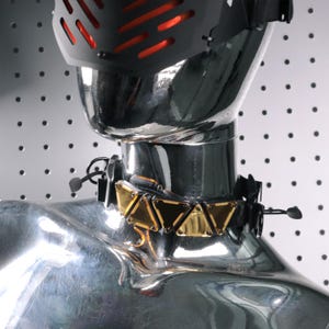 Op de afbeelding: Een zwarte en gouden choker ketting met driehoekige gouden accenten. De ketting staat op een chromen mannequin. Een zwarte helm met rode accenten is zichtbaar boven de ketting.