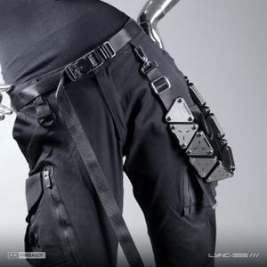 Peut inclure: Pantalon cargo noir doté d'une ceinture tactique avec boucle à dégagement latéral et d'un harnais de jambe amovible. Le harnais présente une série de plaques triangulaires métalliques disposées en rangée verticale. Poches zippées incluses.