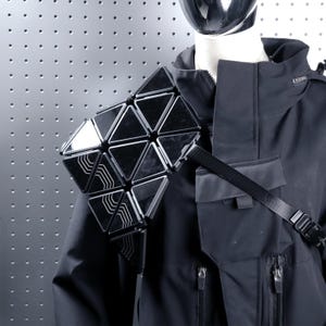 May include: A futuristic, geometric shoulder accessory composed of black triangular panels with reflective surfaces. The accessory is attached to a black jacket with a strap. The design is modern and edgy, suitable for cosplay or avant-garde fashion.