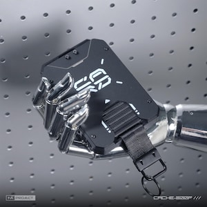 May include: A matte black rectangular wallet with white geometric graphics and the text CACHE-800P. The wallet features a black nylon strap with a metal key ring attachment, secured by visible screws on the front panel surface.