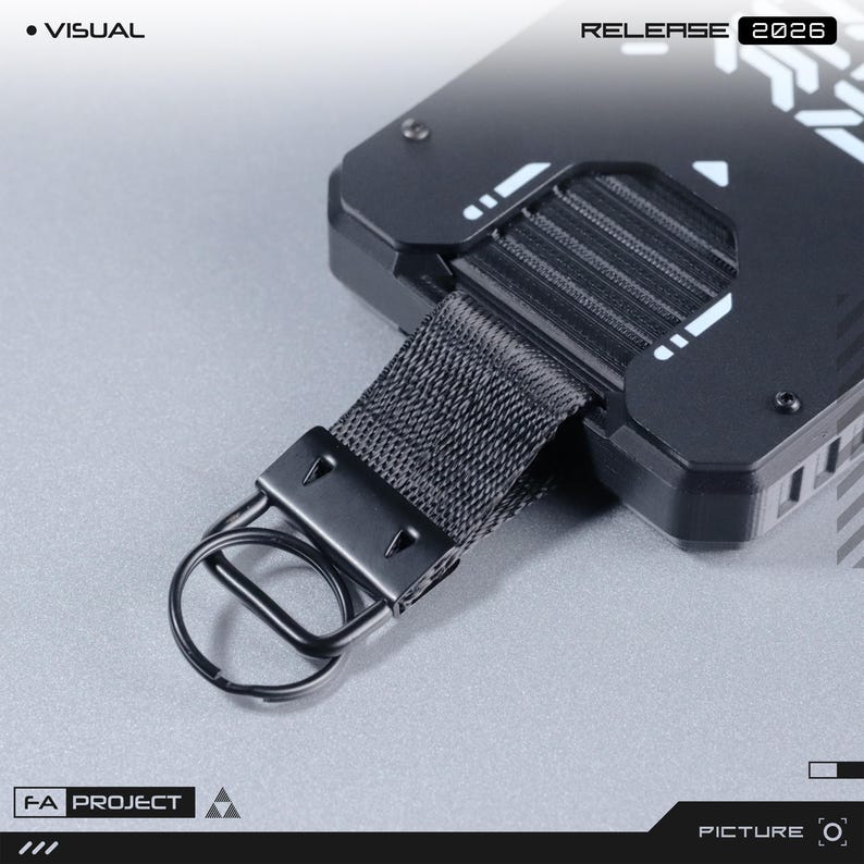May include: A black rectangular device with a matte finish, featuring white geometric accents and the text "VISUAL" and "RELEASE 2026". It includes a black nylon strap attached to a metal split ring, with "FA PROJECT" printed on the lower left corner.