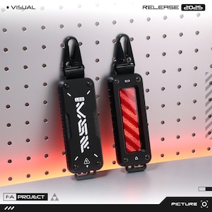 Può includere: Due portachiavi neri dal design futuristico. Un portachiavi ha un fronte nero con testo bianco e l'altro ha una luce LED rossa. Entrambi hanno un moschettone nero e il testo "RELEASE 2025".