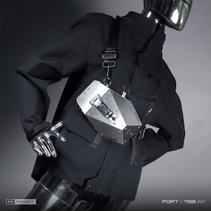 Könnte beinhalten: Eine metallisch silberne, geometrisch geformte Brusttasche mit zentralem Schnallenverschluss und schwarzem verstellbarem Riemen, getragen über einer schwarzen Jacke. Die Tasche trägt den Aufdruck FA PROJECT und PORT - 755 /// im technischen Stil.