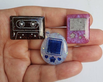 May include: Three resin keychains featuring a cassette tape, a Tamagotchi-style game, and a small rectangle with a star design. The cassette tape is black with red glitter, the game is white with blue and yellow details, and the rectangle is pink with silver glitter.