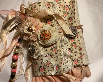 May include: A small, handmade journal covered in a floral fabric with pink and white accents. The journal is decorated with lace, ribbons, and beads.