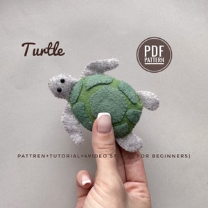 May include: A grey and green felt turtle toy with black eyes. The toy is being held in a hand. The text "Turtle" is written above the toy and "PDF PATTERN" is written in a circle above the hand. The text "PATTREN+TUTORIAL+4VIDEO STITCH (FOR BEGINNERS)" is written below the toy.