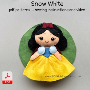 May include: A felt Snow White doll with a yellow dress, blue bodice, and red cape, set on a green felt circle. The doll has black hair, a red bow, and a smiling face. Text at the top reads "Snow White" and "pdf patterns + sewing instructions and video."